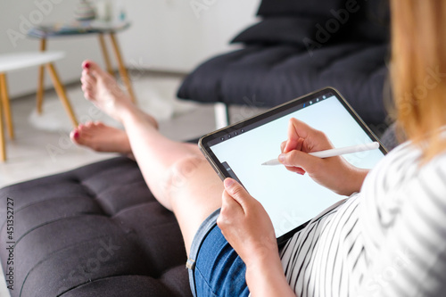 Woman working or drawing on the tablet with pencil and laying on the sofa. A designer or artist works from home