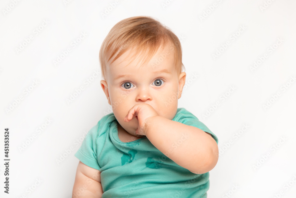 cute Caucasian one year old baby sucks thumb and scratches his first teeth. toddler boy teasing on white background in studio. baby care and child health concept