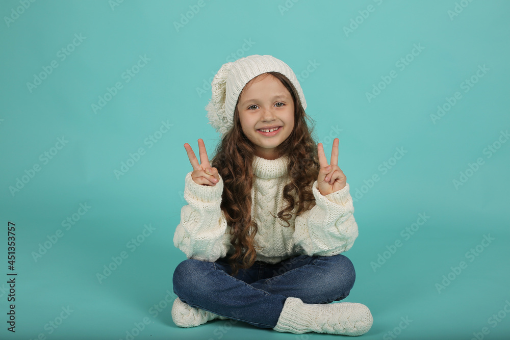 a beautiful happy brown-haired girl with curly hair in a white sweater and a white knitted hat is sitting on a blue background a beautiful happy brown-haired girl with curly hair in a white sweater and a white knitted hat is sitting on a blue background