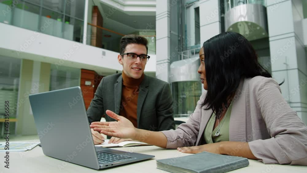 Afro-American businesswoman explaining something on laptop screen to Caucasian male colleague as he taking notes at desk in modern office center Afro-American businesswoman explaining something on laptop screen to Caucasian male colleague as he taking notes at desk in modern office center
