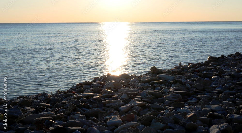 empty rocky beach at sunset with the setting sun and a sunny path on the calm face of the sea, beautiful coastal landscape of a deserted stone coast and calm golden waves