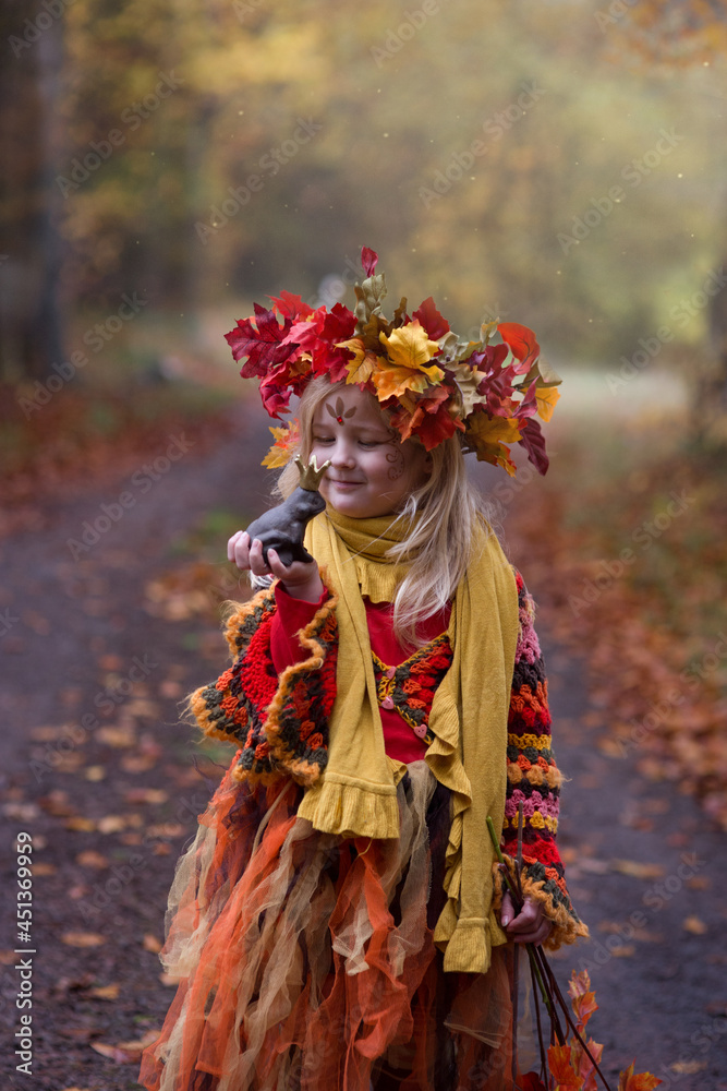 Herbstfee, ein kleines blondes Mädchen mit dem bunten Herbstkranz auf dem Kopf steht im bunten Herbstwald. Das Gesicht ist mit der Herbstmuster geschminkt. In der Hand ist eine Kastanie. Herbstfee, ein kleines blondes Mädchen mit dem bunten Herbstkranz auf dem Kopf steht im bunten Herbstwald. Das Gesicht ist mit der Herbstmuster geschminkt. In der Hand ist eine Kastanie.