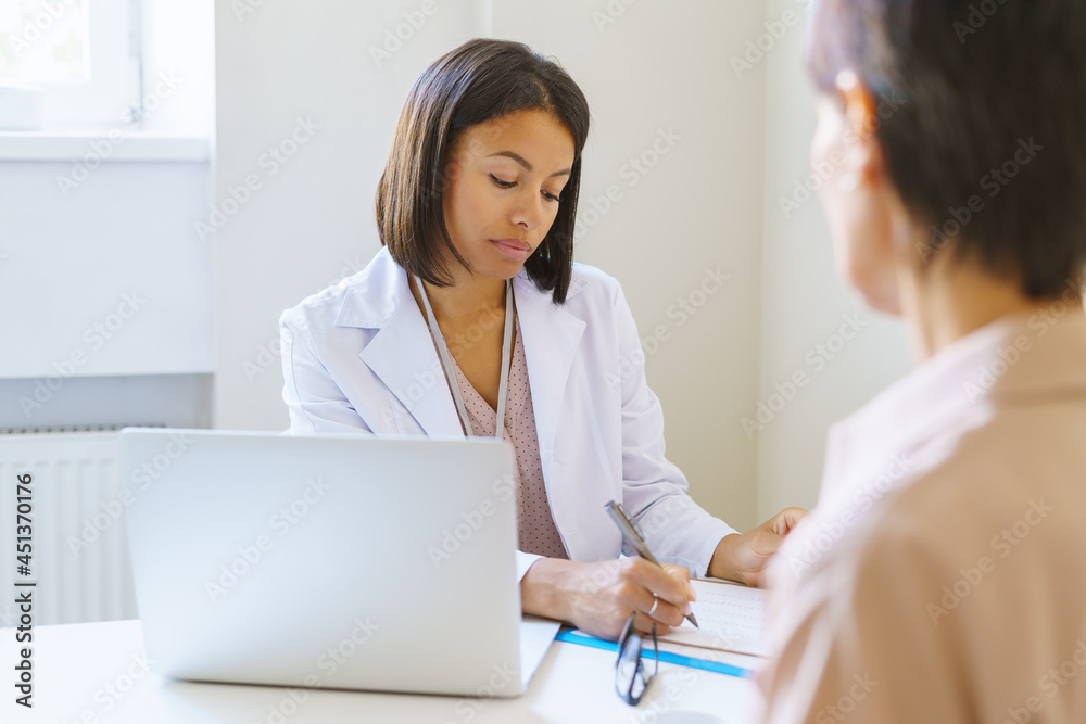 Serious afro american woman doctor in white medical uniform listening female patient, writing symptoms in clipboard, female physician filling medical documents while consulting client in clinic office Serious afro american woman doctor in white medical uniform listening female patient, writing symptoms in clipboard, female physician filling medical documents while consulting client in clinic office