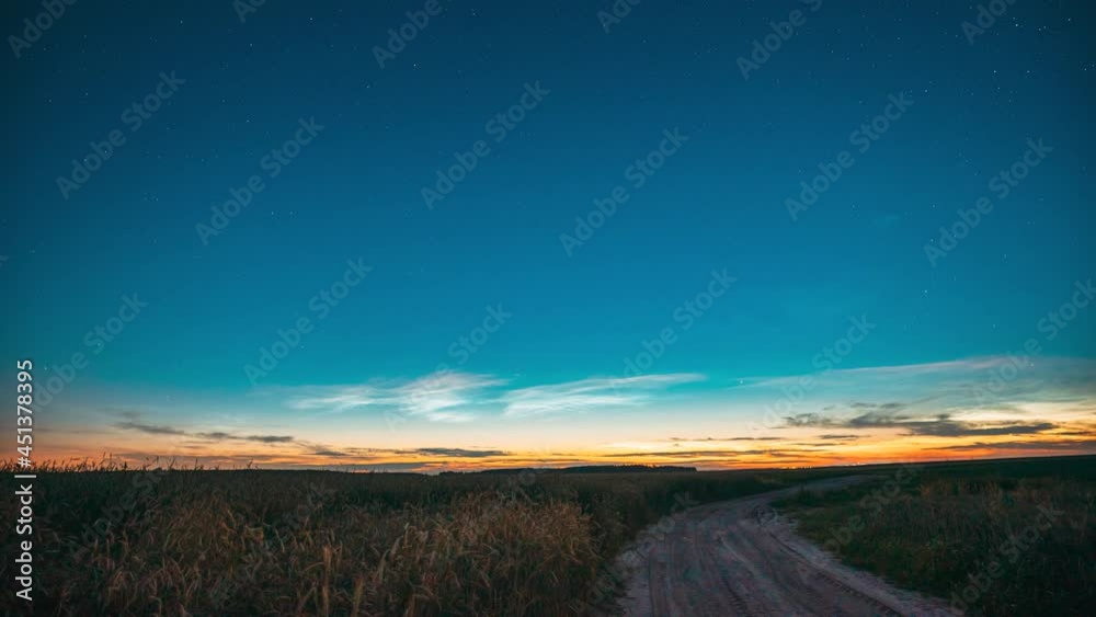 Time Lapse 4K Night Starry Sky With Glowing Stars Above Countryside Landscape With Country Road And Wheat Fields. Noctilucent Clouds Above Rural Wheat Field In Summer. Summertime. TimeLapse Time-Lapse