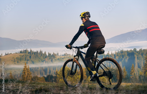 Fototapeta Naklejka Na Ścianę i Meble -  Male cyclist in cycling suit riding bike with coniferous trees and hills on background. Man bicyclist enjoying bicycle ride in mountains. Back view. Concept of sport, biking and active leisure.