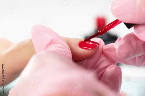 Close up process of applying red varnish. Woman in salon receiving manicure by nail beautician. Manicurist holding client s finger with red nail polish and brush  macro. Shallow depth of field. 
