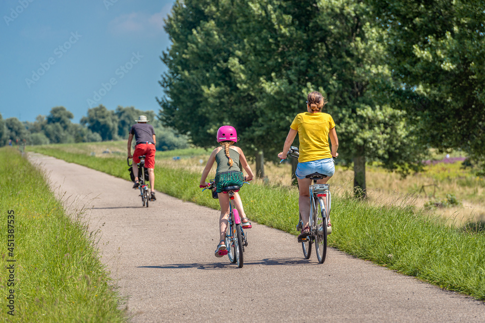 A father, mother and daughter are cycling on a Dutch dike. The little girl is wearing a pink bicycle helmet. It is a sunny day in summertime. The photo was taken in the province of North Brabant.