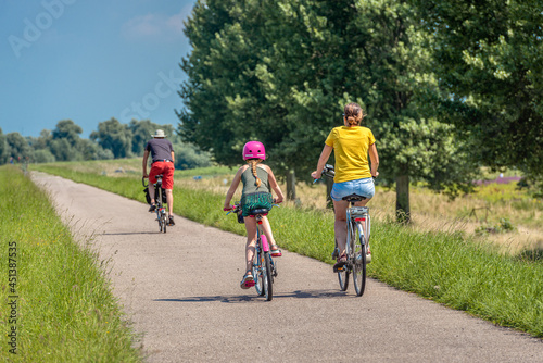 A father, mother and daughter are cycling on a Dutch dike. The little girl is wearing a pink bicycle helmet. It is a sunny day in summertime. The photo was taken in the province of North Brabant.