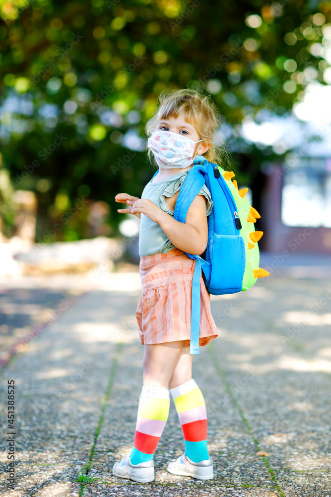 Naklejka premium Little toddler girl on her first day on way to playschool with medical mask against corona covid virus. Healthy beautiful baby walking to nursery preschool and kindergarten. Happy child with backpack