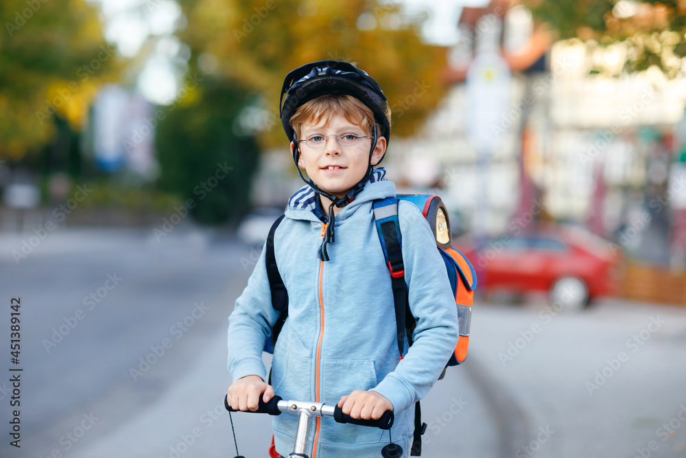 Active school kid boy in safety helmet riding with his scooter in the city with backpack on sunny day. Happy child in colorful clothes biking on way to school.
