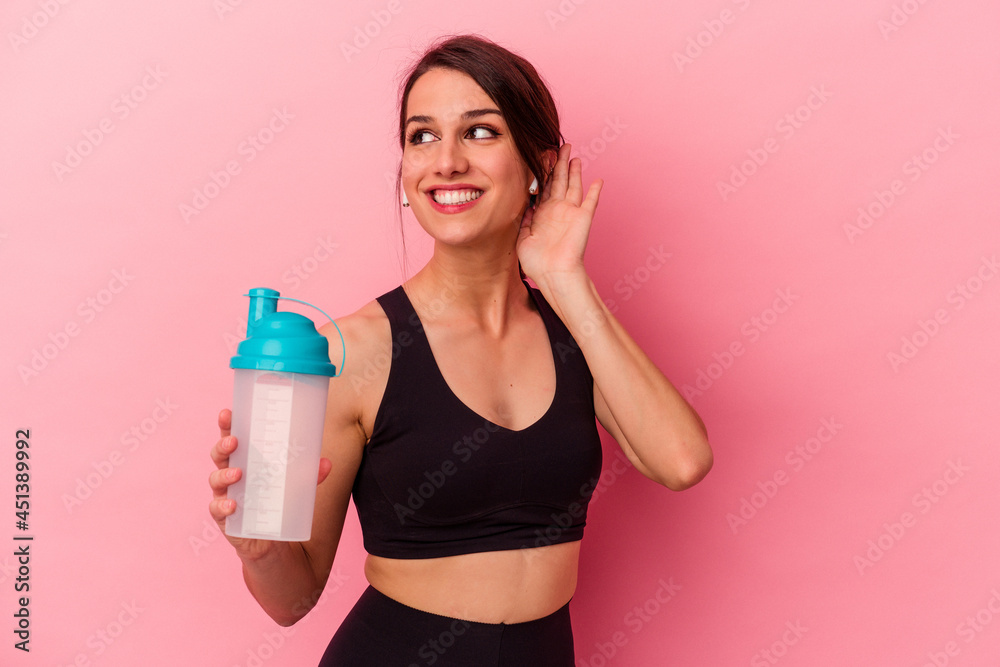 Young caucasian woman drinking a protein shake isolated on pink background trying to listening a gossip. Young caucasian woman drinking a protein shake isolated on pink background trying to listening a gossip.