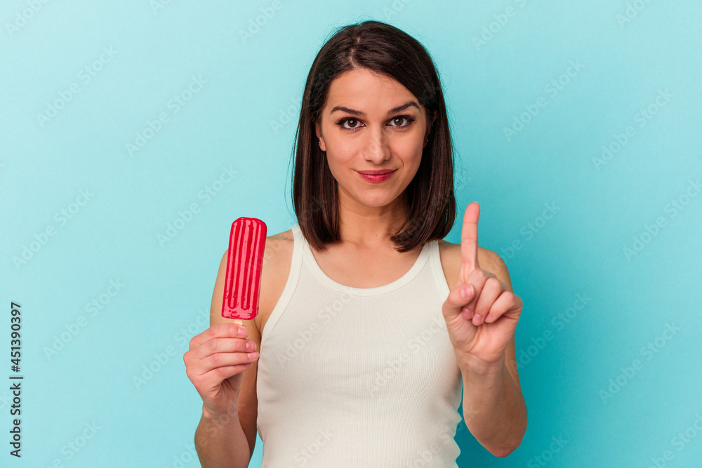 Young caucasian woman holding an ice cream isolated on blue background showing number one with finger. Young caucasian woman holding an ice cream isolated on blue background showing number one with finger.