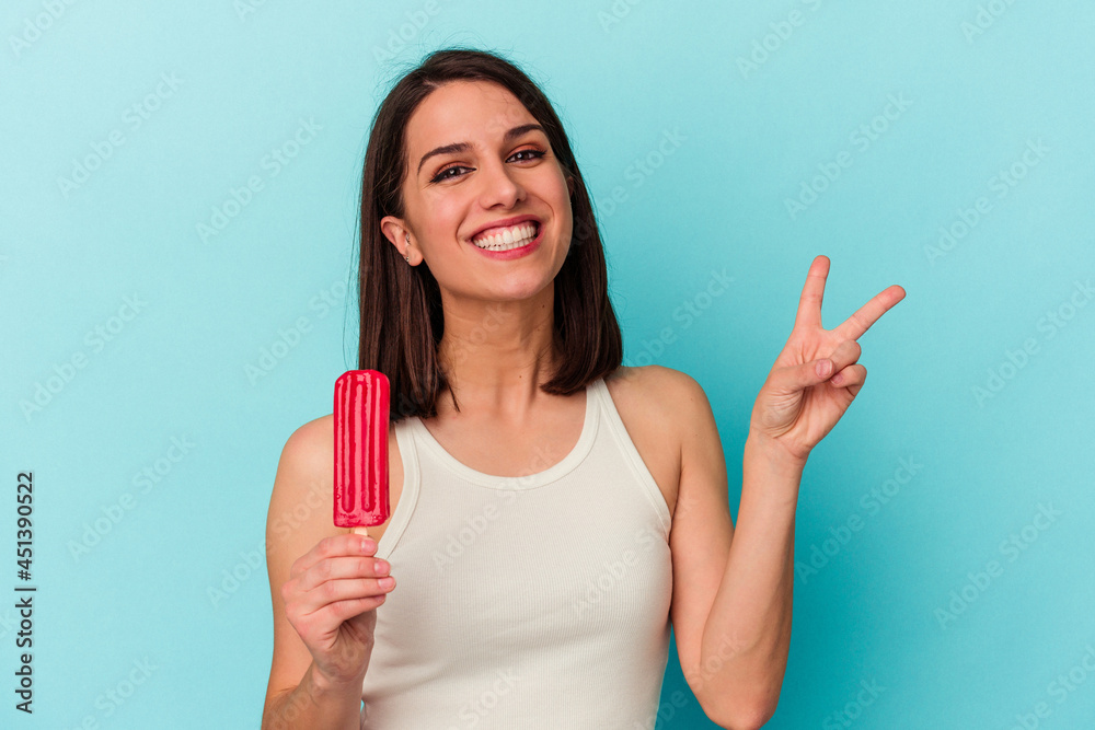 Young caucasian woman holding an ice cream isolated on blue background joyful and carefree showing a peace symbol with fingers. Young caucasian woman holding an ice cream isolated on blue background joyful and carefree showing a peace symbol with fingers.