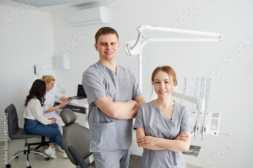 Two dentists - a man and a woman are standing in the office  and in the background sits a patient and an assistant. Dentist s office