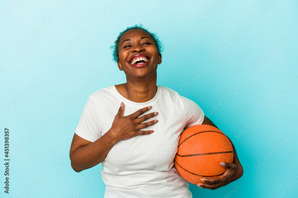 Young african american woman playing basketball isolated on blue background laughs out loudly keeping hand on chest.