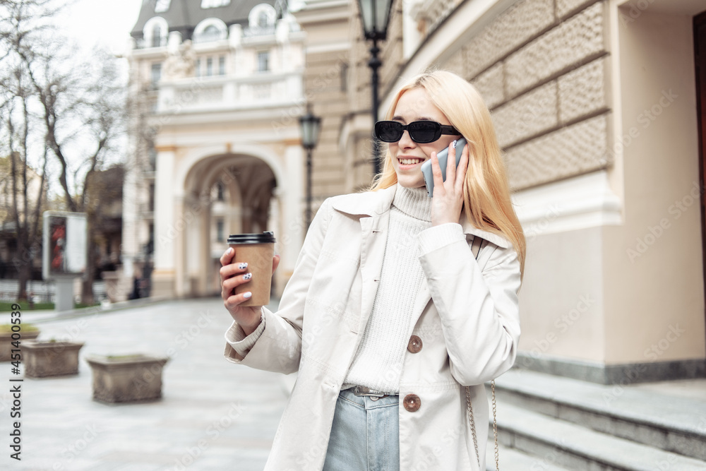 Young blonde woman in sunglasses talking on the phone and holding a cup of coffee in city Young blonde woman in sunglasses talking on the phone and holding a cup of coffee in city