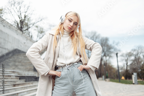 Young stylish blonde woman in trench coat listens to music on headphones outdoor