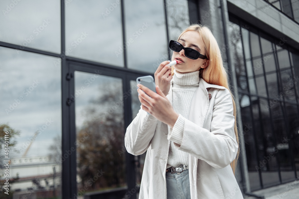 Young stylish fashion woman corrects makeup, paints lips with lipstick near business center