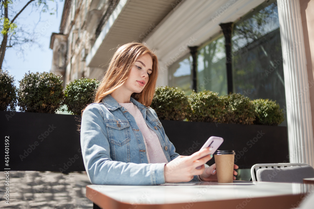 Young beautiful woman using smartphone and drinking coffee while sitting at a table in outdoor cafe