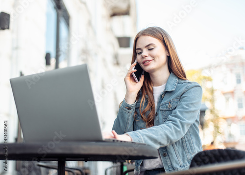 Young beautiful woman freelancer using laptop and talking on phone while sitting at table in outdoor cafe