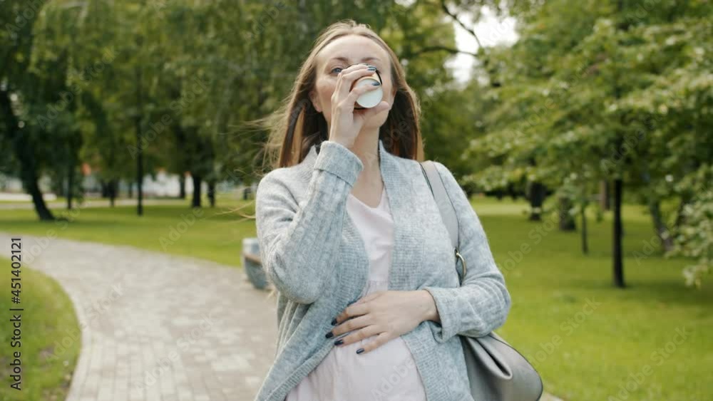 Dolly shot of attractive pregnant lady walking in summer park drinking take away coffee smiling enjoying nature alone with baby. Leisure time and pregnancy concept. Dolly shot of attractive pregnant lady walking in summer park drinking take away coffee smiling enjoying nature alone with baby. Leisure time and pregnancy concept.