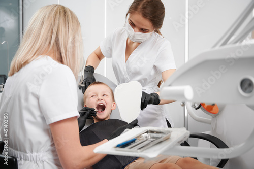 Adorable little boy sitting in dental chair while two female dentists checking kid teeth. Dentist examining little boy teeth with dental instrument. Concept of pediatric dentistry and dental care.