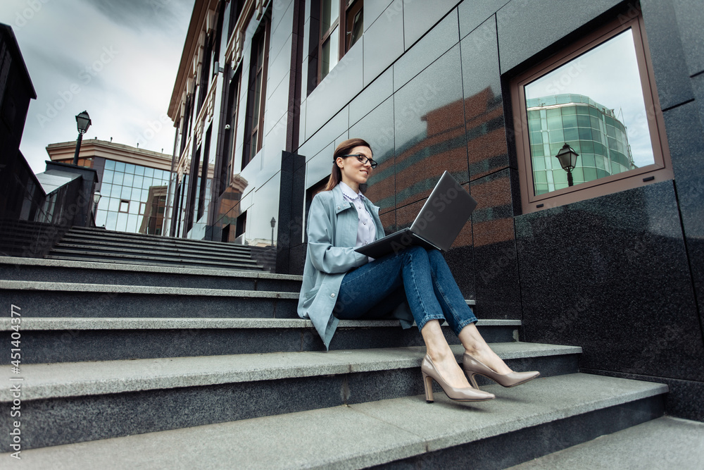 Cute Caucasian woman is using laptop while sitting on stairs outdoors. Distant work. Modern business woman. Uses gadgets. Lifestyle