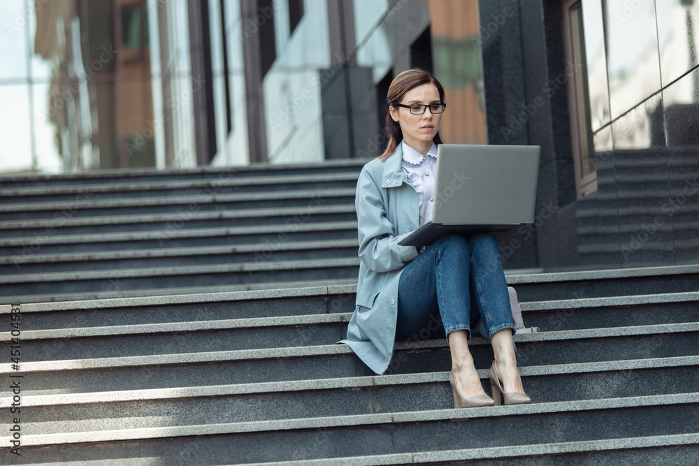 Cute Caucasian woman is using laptop while sitting on stairs outdoors. Distant work. Modern business woman. Uses gadgets. Lifestyle Cute Caucasian woman is using laptop while sitting on stairs outdoors. Distant work. Modern business woman. Uses gadgets. Lifestyle