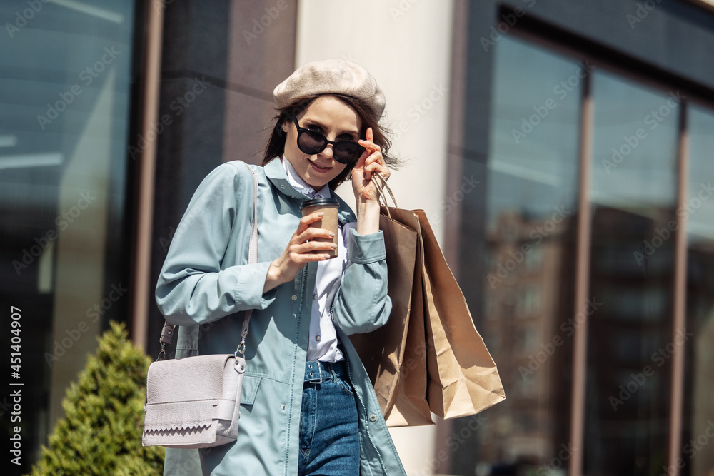 Stylish shopaholic woman in trendy trench coat, beret, sunglasses in city with shopping bags and cup of coffee. Shopping concept. Lifestyle