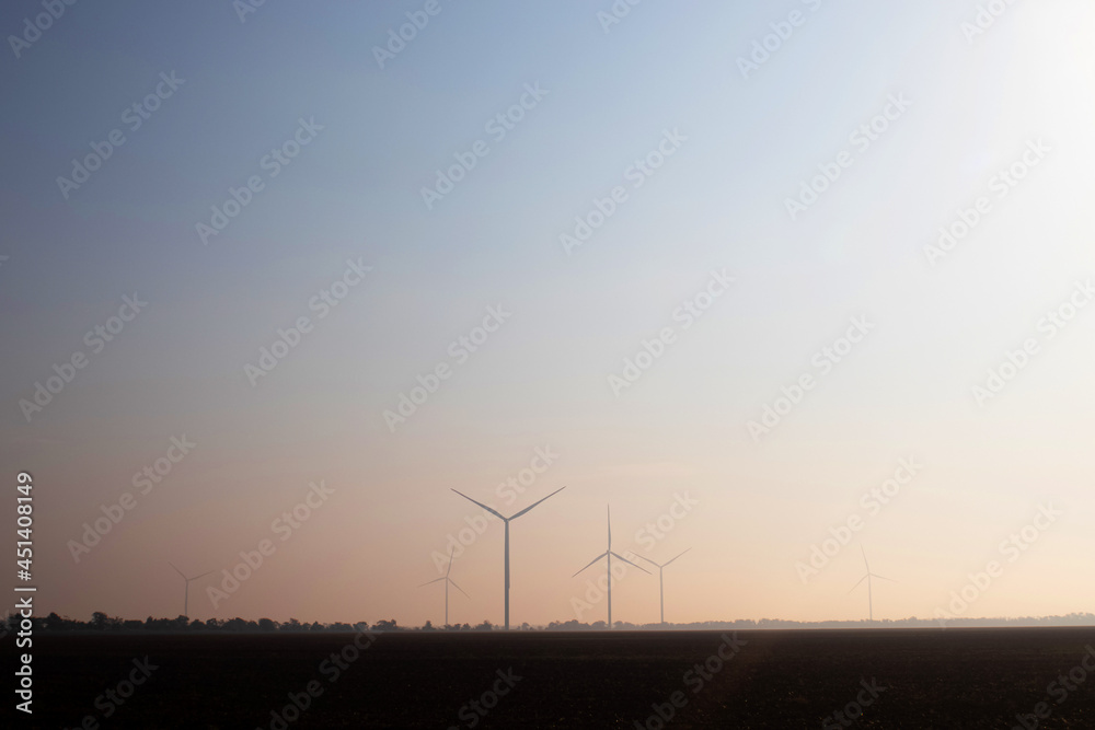 Windmills in the fog near a field of sunflowers. Early morning. See the movement of the blades, as a strong wind is blowing. Wind turbine are installed at different distances and hidden in the fog