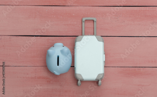 Flat lay vacation holiday and travel planing concept. Mini plastic travel suitcase and piggy bank on red wooden background. Top view