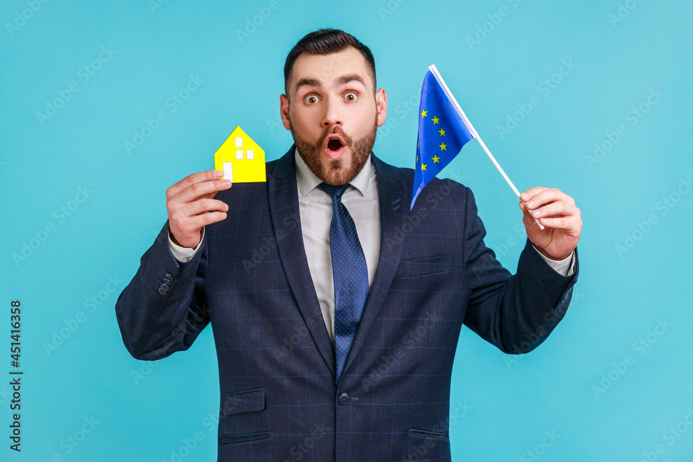 Amazed man with beard wearing official style suit holding European flag and paper house, looking at camera with surprised expression. Indoor studio shot isolated on blue background. Amazed man with beard wearing official style suit holding European flag and paper house, looking at camera with surprised expression. Indoor studio shot isolated on blue background.
