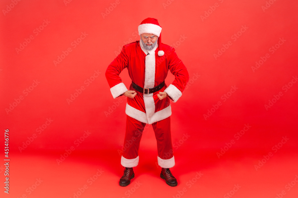 I am strong. Portrait of funny elderly man with gray beard wearing santa claus costume standing looking at camera with proud face, demonstrating power. Indoor studio shot isolated on red background.