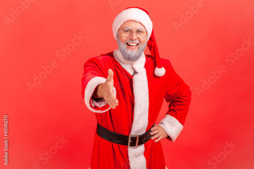 Smiling friendly elderly man with gray beard wearing santa claus costume giving hand for handshaking, welcoming people to Christmas party. Indoor studio shot isolated on red background.