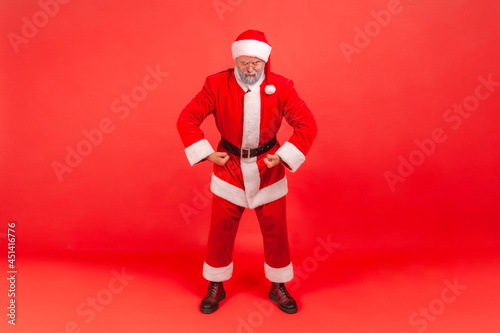 I am strong. Portrait of funny elderly man with gray beard wearing santa claus costume standing looking at camera with proud face, demonstrating power. Indoor studio shot isolated on red background.