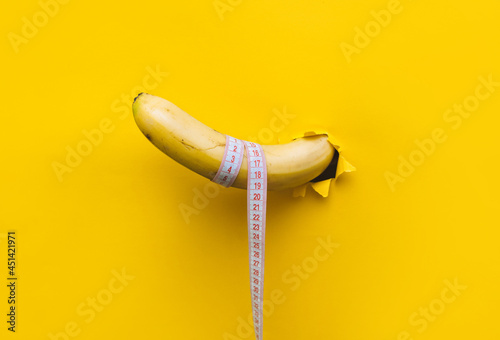 A banana with a tape measure wrapped around it appears through a torn hole in yellow paper. The concept of healthy food, diet and potency. Copy space. photo