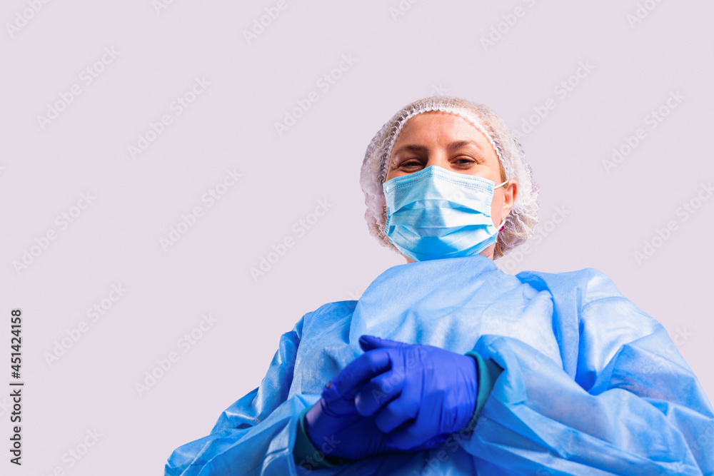 Photo from below of a senior female doctor in a medical gown, face mask and protective blue gloves looking down into the camera with smiling eyes. Photo from below of a senior female doctor in a medical gown, face mask and protective blue gloves looking down into the camera with smiling eyes.