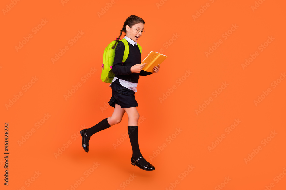 Full length body size view of pretty cheerful schoolgirl jumping reading diary isolated over bright orange color background