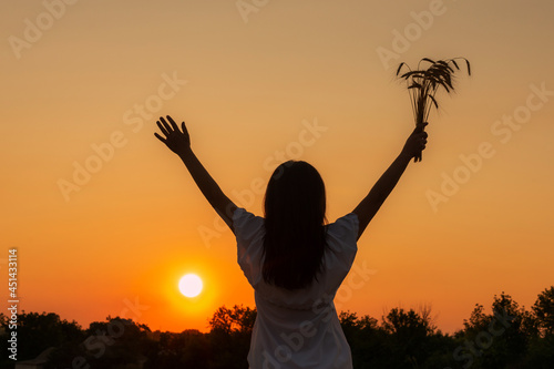 Happy woman raising hands over sunset sky, enjoying life and nature. Amazing girl hold wheat ears in hand. Silhouette of girl in sunlight rays. Freedom and feel good concept