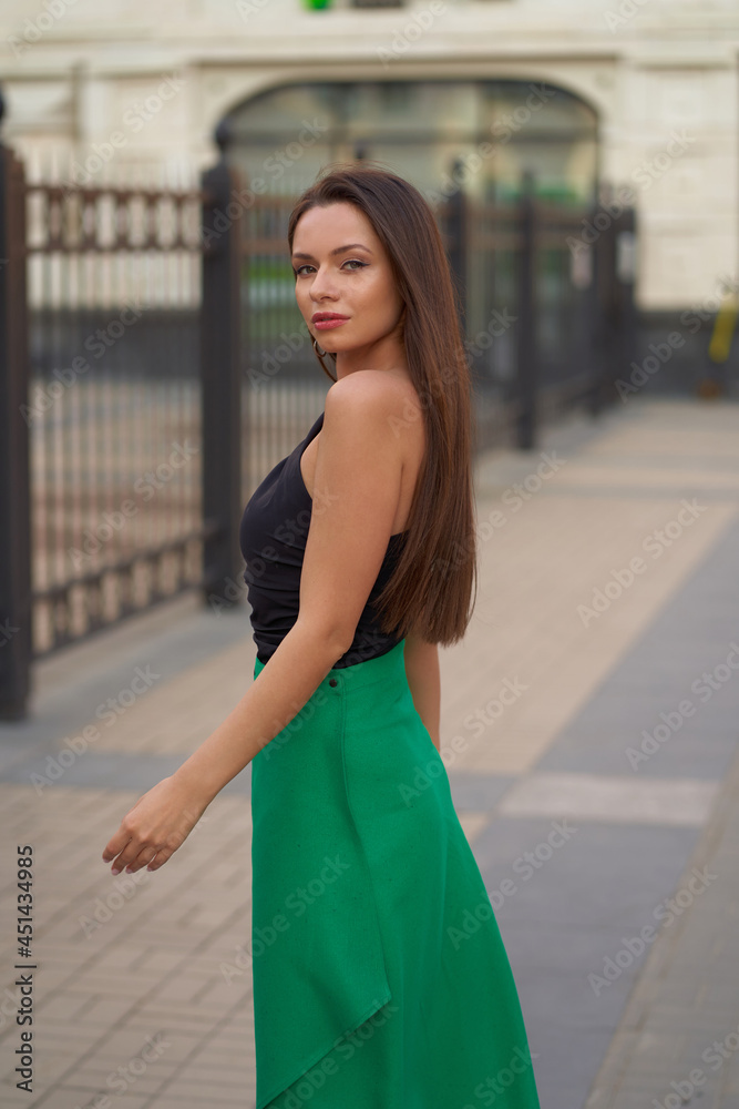Elegant woman in black top and green skirt. Pretty girl with long straight brunette hair in black sunglasses. Female model walking at city street
