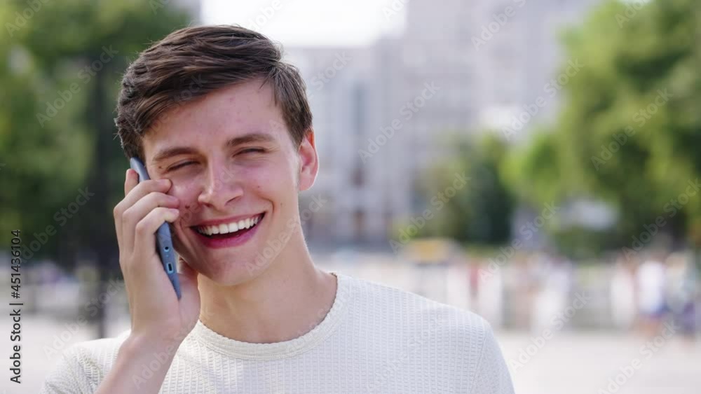 Young man in casual wear talking on phone and smiling, blurred buildings and trees on background. Closeup friendly conversation in city on bright summer day. Concept of communication Young man in casual wear talking on phone and smiling, blurred buildings and trees on background. Closeup friendly conversation in city on bright summer day. Concept of communication
