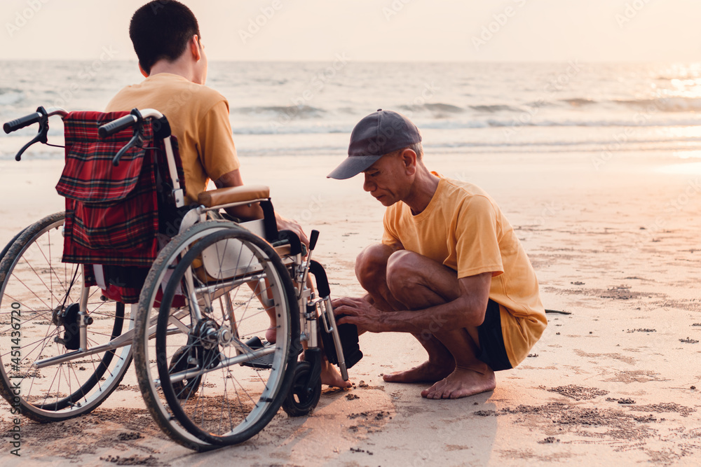 Happy disabled teenage boy on wheelchair playing and relaxing with parent, Activity outdoors with father on the beach background, People having fun and diverse family concept.
