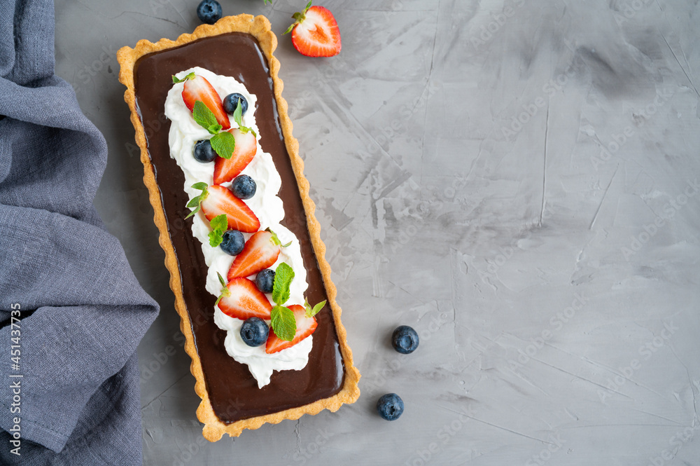 Chocolate cream tart with whipped cream, fresh blueberries and strawberries on top on a dark background. Summer dessert.
