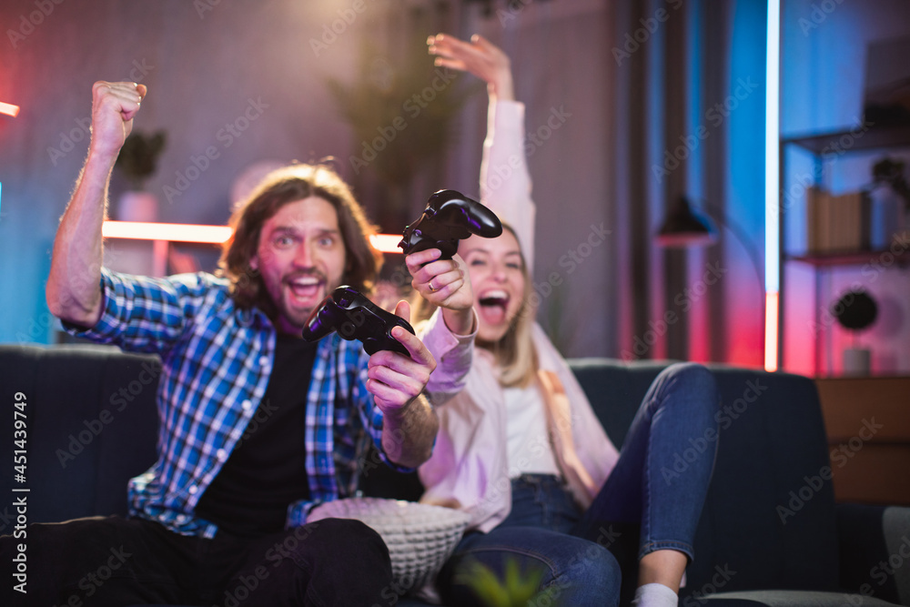 Cheerful young woman with man enjoying winning in video games while sitting on couch. Caucasian couple in casual outfit spending free time with fun at home.
