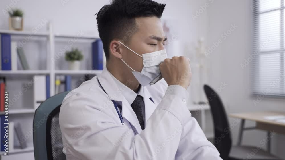 slow motion with headshot sick asian man doctor holding a pen in face mask is coughing into his fist while sitting working in a white hospital office. slow motion with headshot sick asian man doctor holding a pen in face mask is coughing into his fist while sitting working in a white hospital office.