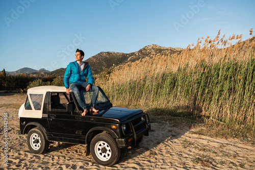 Young Man sits on the roof of his off-road car watching the sunset in a wild beach after a day of travel and adventure