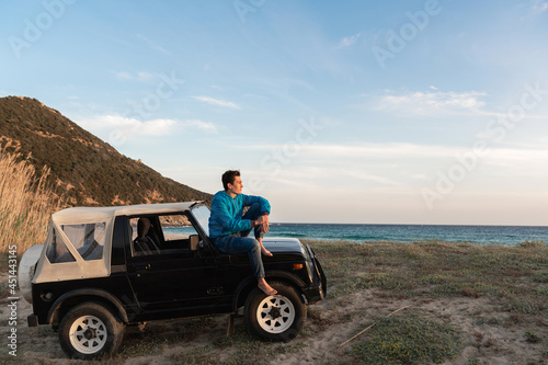 Young man sitting on his off-road car parked in front of a wild beach enjoying and relaxing looking the view