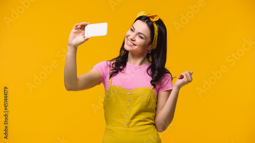 Smiling woman in headband taking selfie on smartphone isolated on yellow.
