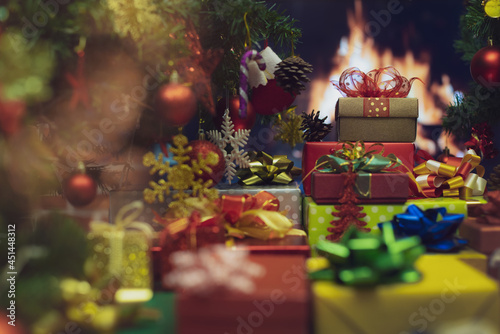Pile of shiny luxury colorful gift boxes. Christmas and new year celebrate gifts stack beside full decorated Xmas tree with flame of fire in fireplace blur in background to give for family members © Sura Nualpradid