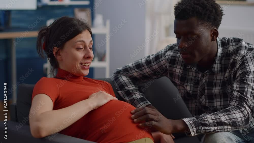 Close up of interracial couple with pregnancy at home holding hands on belly. Mixed race young partners expecting child and touching baby bump while sitting on living room couch Close up of interracial couple with pregnancy at home holding hands on belly. Mixed race young partners expecting child and touching baby bump while sitting on living room couch
